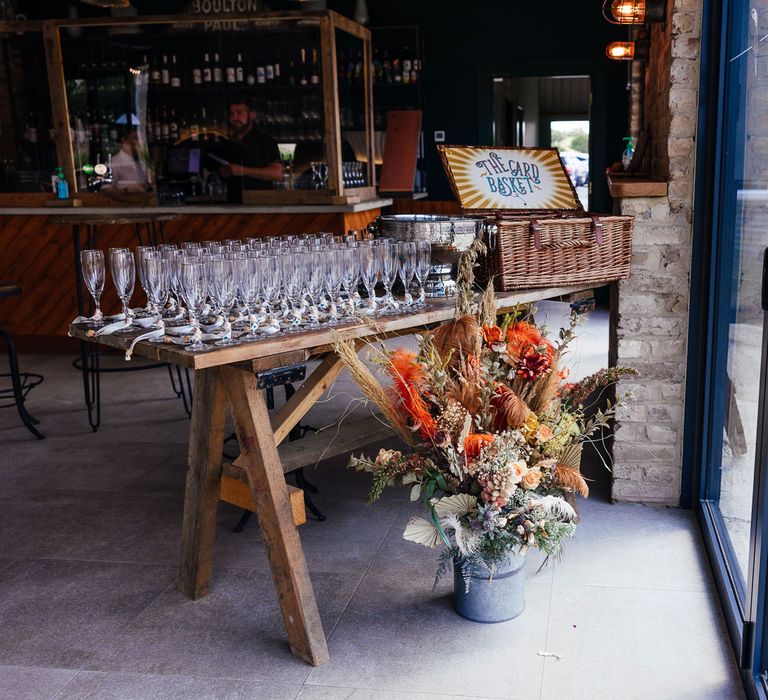 Rustic wooden table with glasses and picnic style wedding card box with milk churn full of mixed dried florals for festival inspired wedding