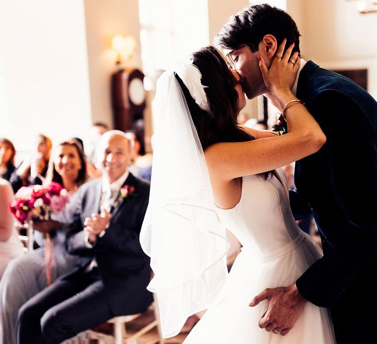 Bride in tulle wedding dress, veil and feathered headband kisses groom in dark suit during wedding ceremony in the Cotswolds