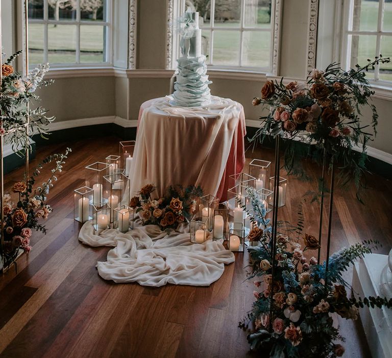 Light blue ruffle wedding cake on a blush pink table cloth surrounded by pilar candles in hurricane vases at Newburgh Priory