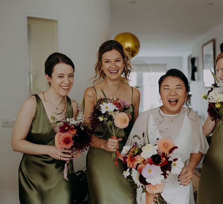 Bride laughs with her bridesmaids as they hold bright floral bouquets and bridesmaids wearing olive green satin dresses  | Irene Yap Photography