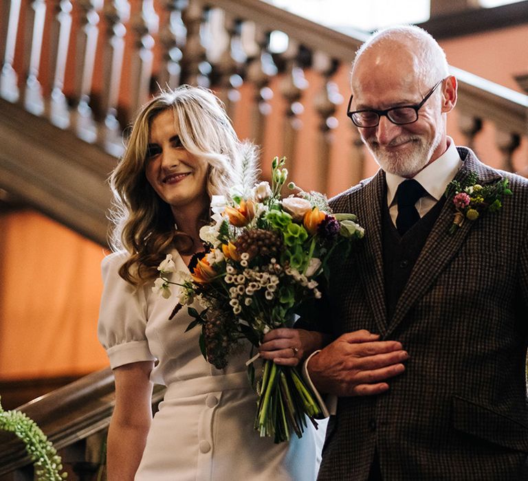 Father of the bride in checkered suit walks the bride downstairs in puff sleeve wedding dress and colourful bouquet