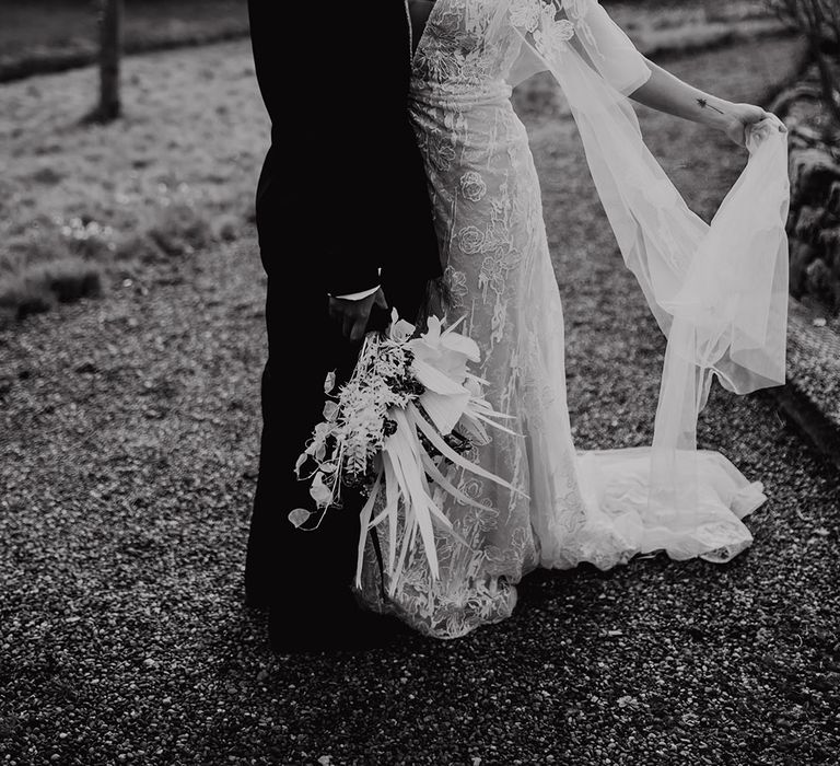 Bride in a lace wedding dress with lunging neckline and watteau train embracing her partner in a black suit 