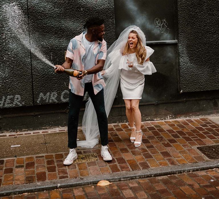 Groom in a pastel patterned shirt and Converse trainers popping champagne with his bride in a short wedding dress and long veil