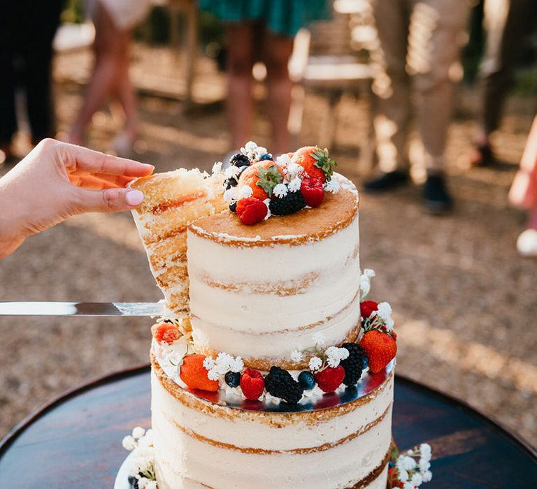 Bride takes a slice of cake from two-tier semi-naked white frosted wedding cake with summer berries and gypsophila 