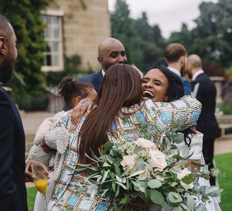 Laughing bride hugs guest in sparkly blue and gold blazer jacket with white rose and green foliage wedding bouquet