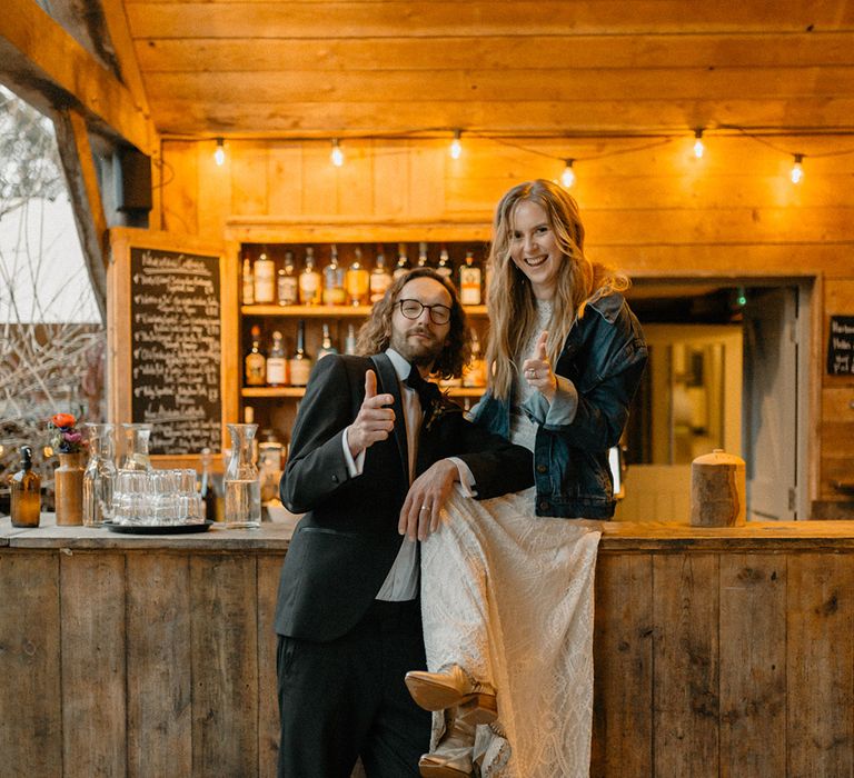 Bride wearing embroidered denim jacket and groom seated at the bar with thumbs up for the camera