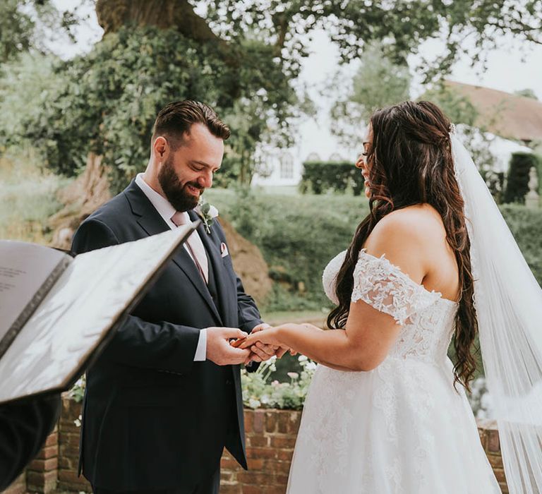 Groom in black suit and pale pink tie puts on brides ring with long brunette hair and off the shoulder lace wedding dress