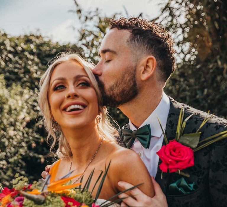 Groom patterned suit jacket and dark green bow tie kisses bride on the head holding bright and colourful bouquet 