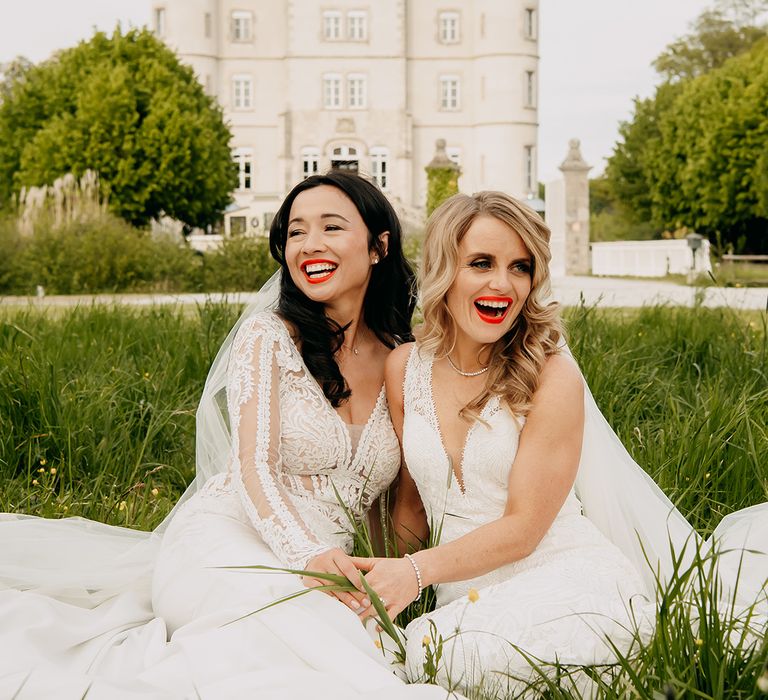 Brides laugh together as they sit in the grass outside their wedding venue 