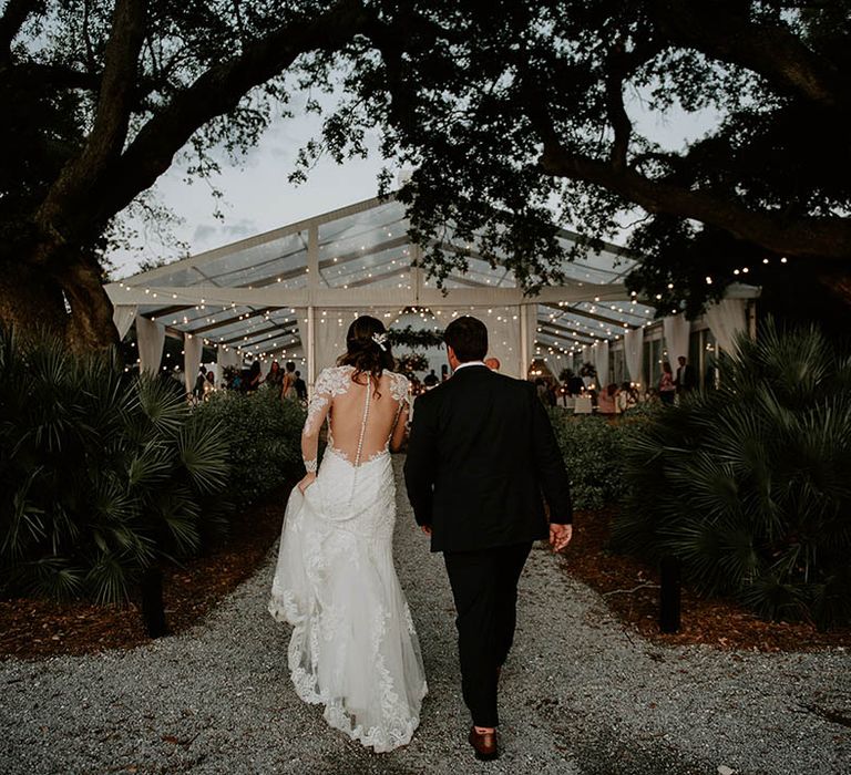 Groom in black suit and bride in button detail illusion back wedding dress walk into their wedding reception 