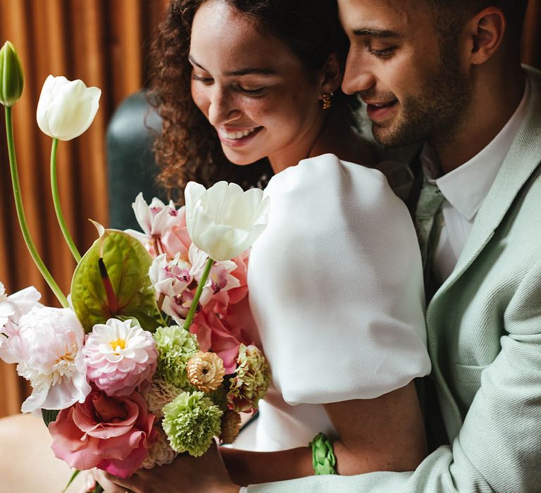 Groom in a pale green suit embracing his bride in a short wedding dress with puffy sleeves holding a pink, green and white spring wedding bouquet 