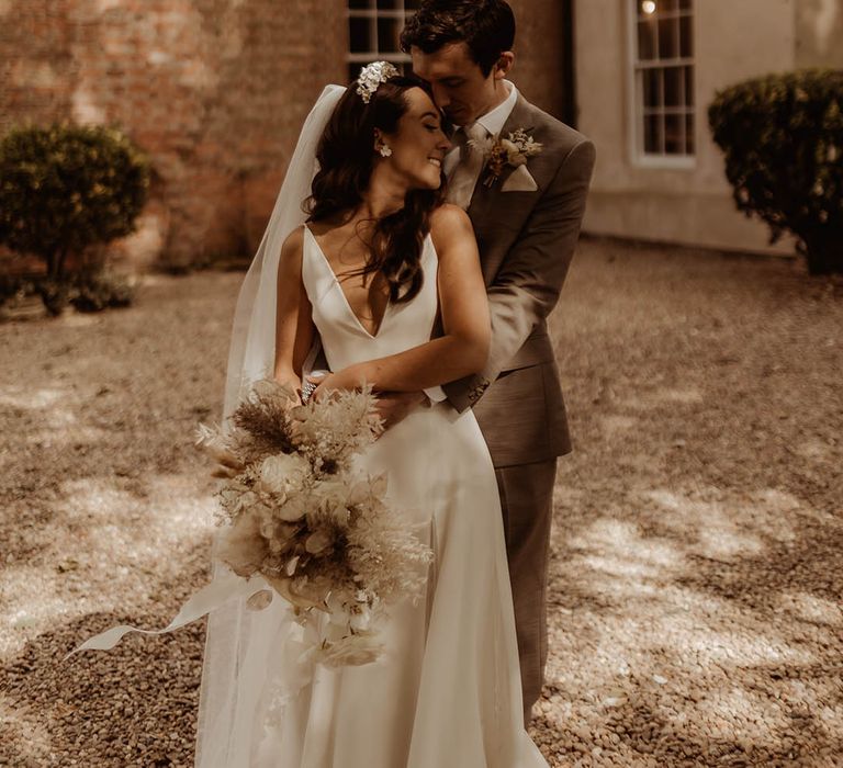 Bride in v-neck wedding dress from Alexandra Grecco is embraced by the groom from behind 