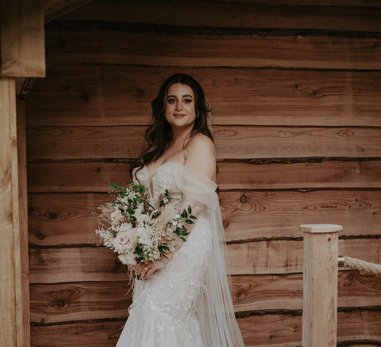 Bride in sparkly wedding dress with tulle skirt and detachable sleeves holding pink and white flower bouquet with dried grass