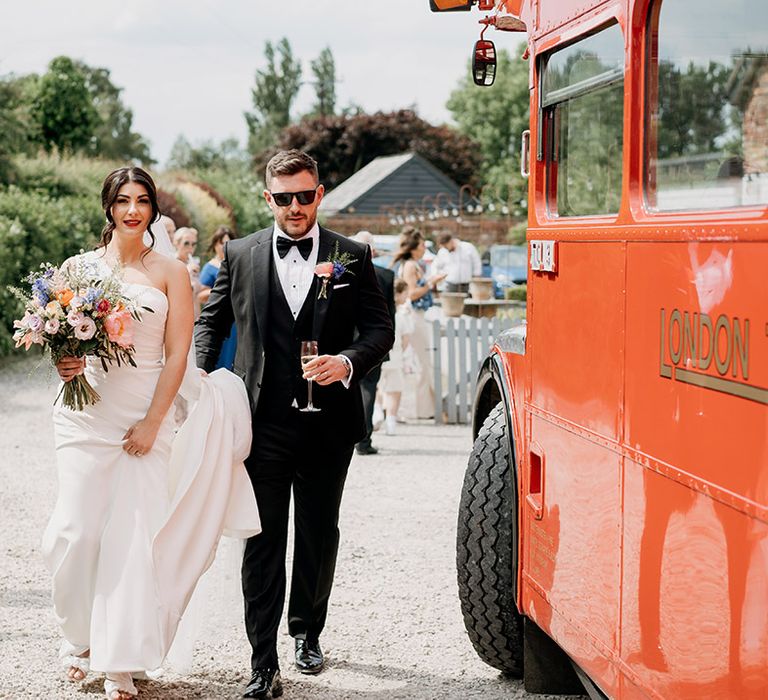 Groom in black tie and sunglasses walks with bride in one shoulder wedding dress next to their bus wedding transport 