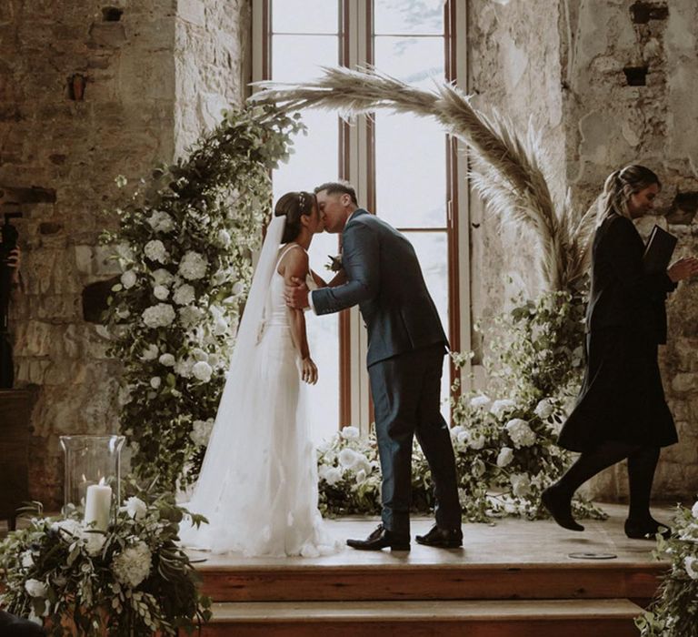 White flower and foliage wedding moongate with pampas grass as the bride and groom share a kiss 