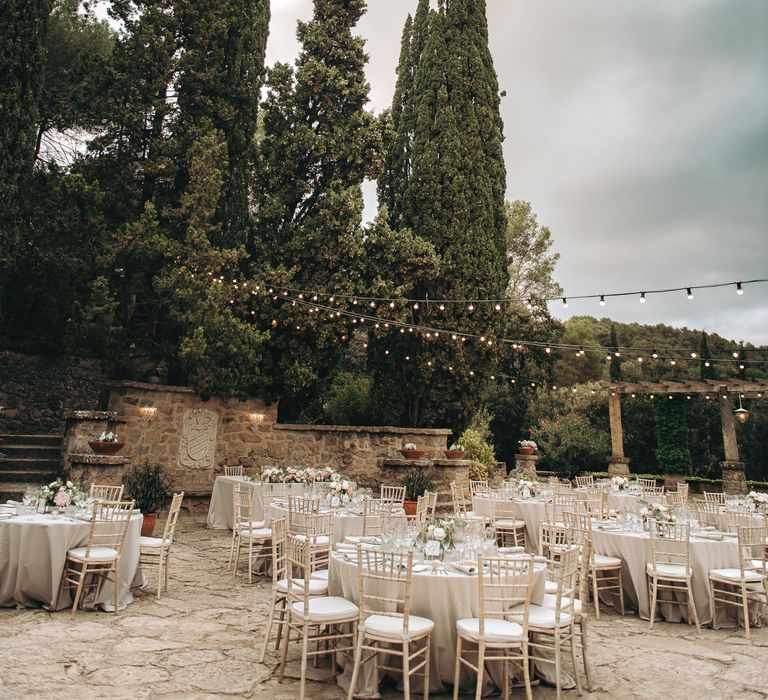 Festoon lighting hangs above round tables for outdoor wedding reception in Barcelona complete with pastel bouquets and rustic feel