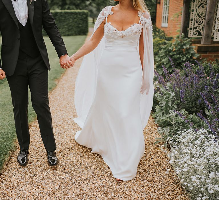 Groom in black tie walks around the venue with the bride in a handmade wedding dress with bridal cape 