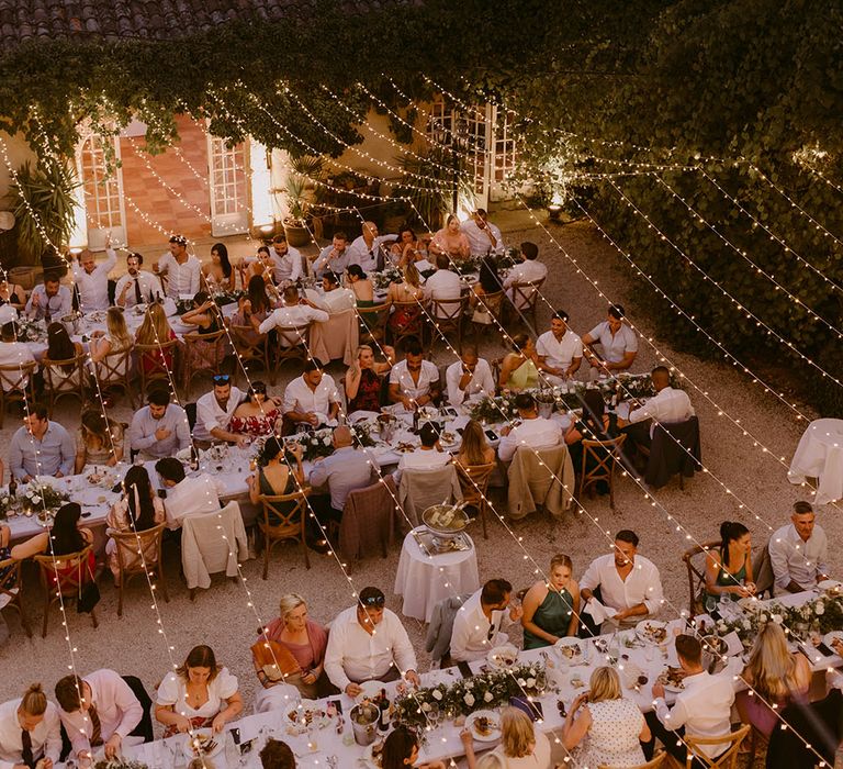 Wedding guests sit at banquet tables during outdoor wedding reception with fairy light canopy hanging above 