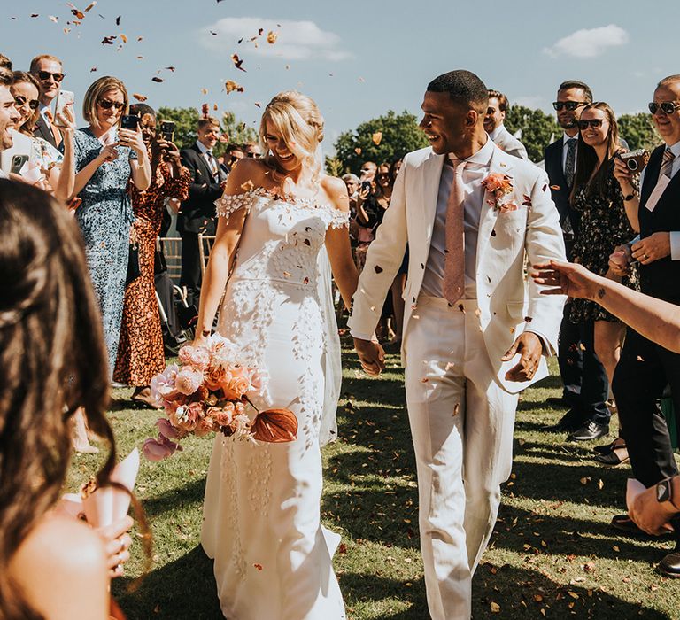 Bride in a leaf design off the shoulder wedding dress has confetti moment with groom in a cream suit and pink tie 