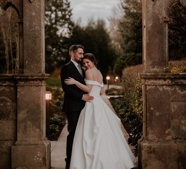 Groom in a blue suit hugs the bride in a button back wedding dress underneath an arch 