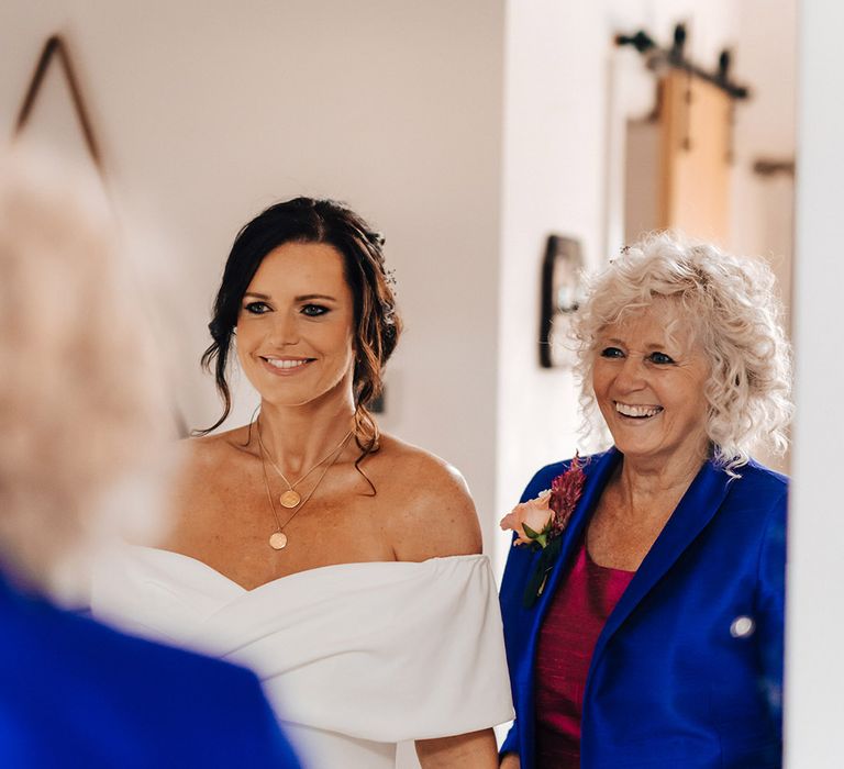 Bride looks in the mirror at her final bridal look with her mother in a bright blue blazer