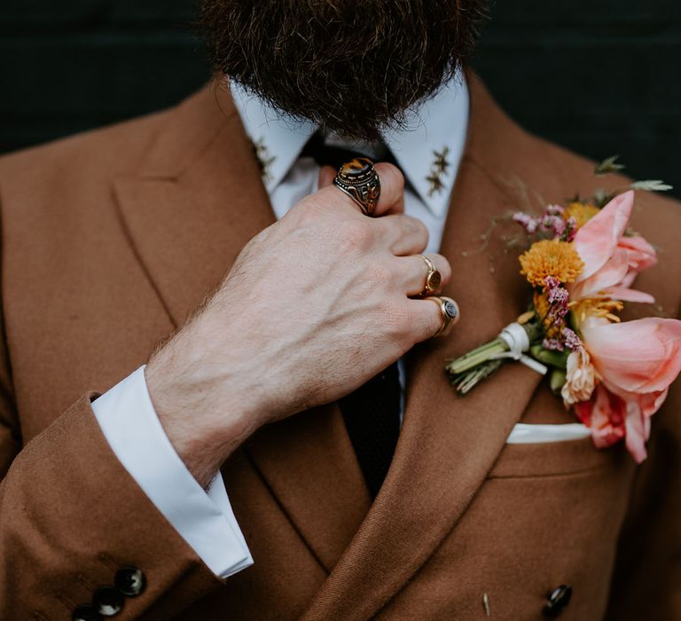 Groom wears brown retro suit complete with pink floral buttonhole and wears silver statement rings