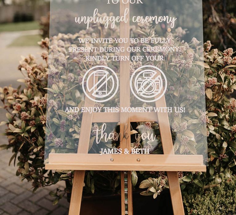 Clear acrylic wedding welcome sign on a wooden easel with pink flowers and foliage 