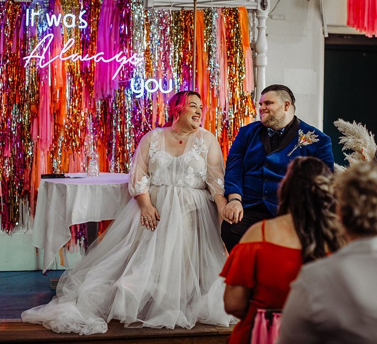 Bride in corset top wedding dress with leaf detailed lace overlay, pink stitching around the waist and red gemstone necklace holding hands with groom and in midnight blue blazer with galaxy patterned bowtie and autumnal boutonniere sitting at the alter