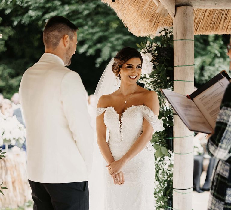 Bride in floral lace wedding dress standing at the outdoor ceremony area with the groom in a white tuxedo 