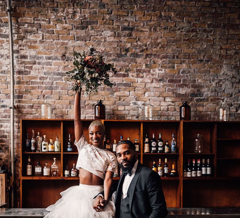 Bride holds her eucalyptus bridal bouquet in the air whilst sitting on the bar next to the Groom at industrial wedding venue