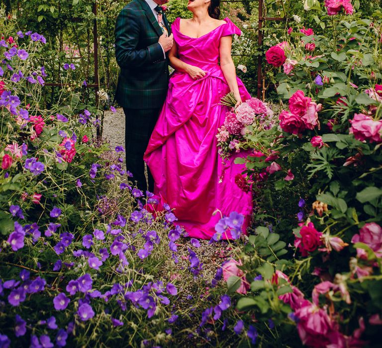 Bride in off the shoulder front ruching pink wedding dress laughing with groom in dark forest green tartan suit with patterned tie, white pocket square and pink and green boutonniere