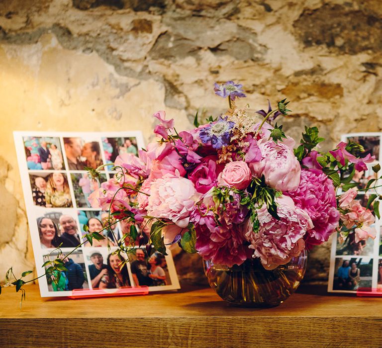 Light pink roses, green foliage, lavender peonies and varying pink peonies floral arrangement in spherical opaque vase with large photo collages on either side