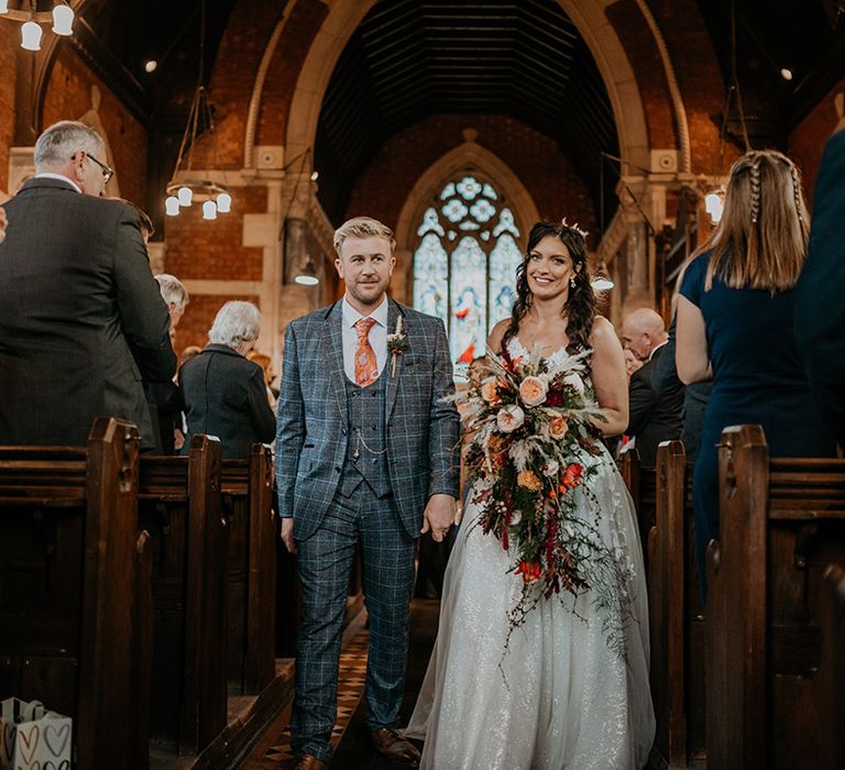 The groom in a three piece grey checkered suit and orange tie walks back down the aisle as a married couple with the bride in a lace and tulle princess skirt wedding dress