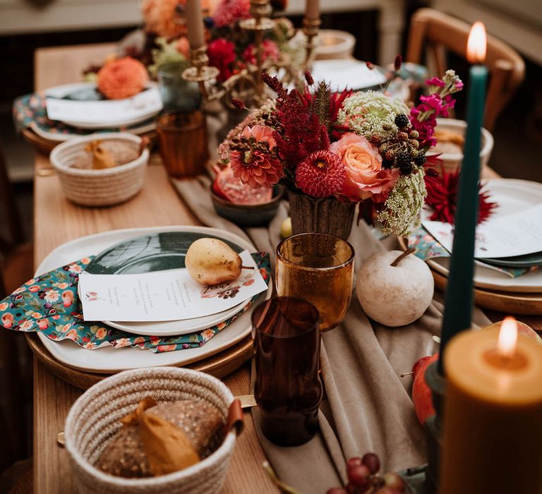 Red, brown and dark green tapered candles in gold candlesticks, tall orange pillar candles, autumnal dried flowers, seasonal fruit and vegetables, and hand-painted silk napkins 