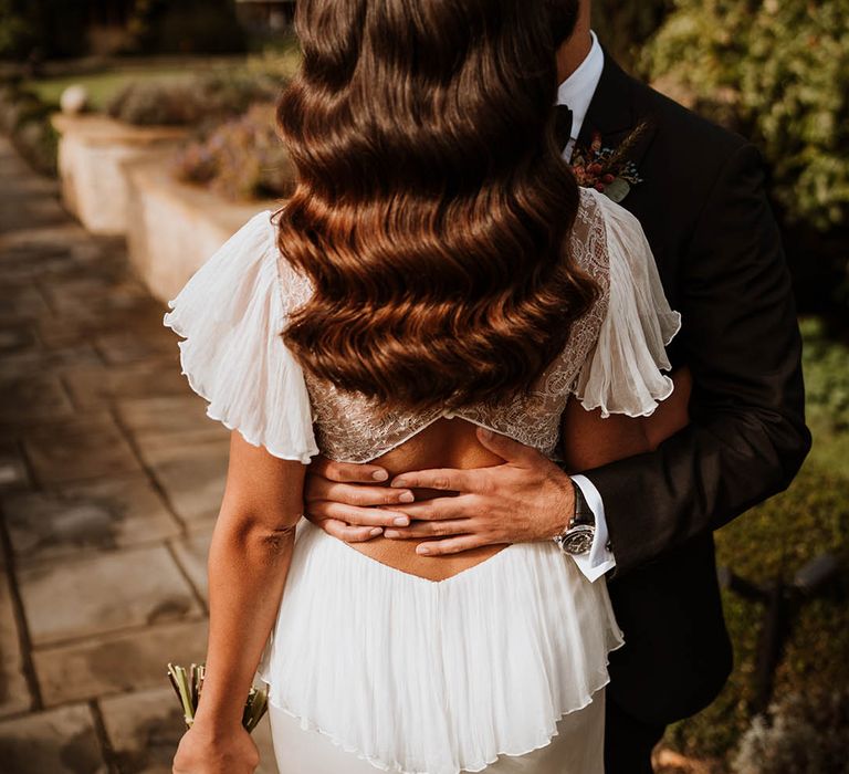 Bride wearing lace open back wedding dress with butterfly sleeves holding autumnal dried flower bouquet and groom in classic black tuxedo embracing her