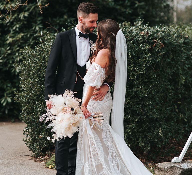 Bride with waved brown hair in a half uo half down style with veil in a boho strapless gown gazes into the groom's eyes who wears a black tux 