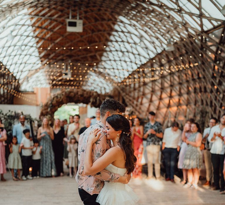 Bride with brown hair in a half up half down style in a ruffle wedding dress doing the first dance with the groom in a pink summer shirt at The Gridshell