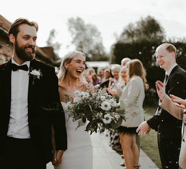 Groom in black tie with a white flower buttonhole exits from the ceremony to confetti with the bride in a fitted wedding dress with spaghetti straps with white anemone bouquet 