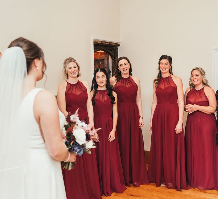 Bridesmaids in burgundy illusion neck dresses smiling as they get the first look at the bride in her wedding dress 