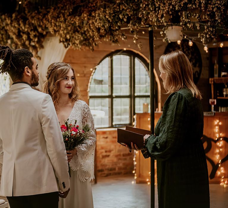 Bride in boho long sleeve lace wedding dress with tassels and groom in white blazer standing at the aisle of barn wedding venue with celebrant 