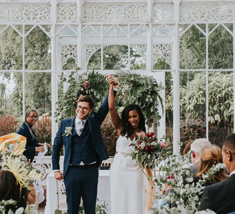 Bride in satin wedding dress and room in blue three piece suit holding hands at The Horniman Museum glasshouse venue with eucalyptus aisle flowers 