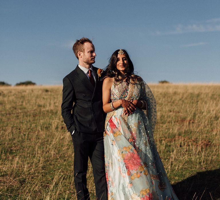 Bride in colourful lehenga leans into her groom during couples portraits in fields outdoors 