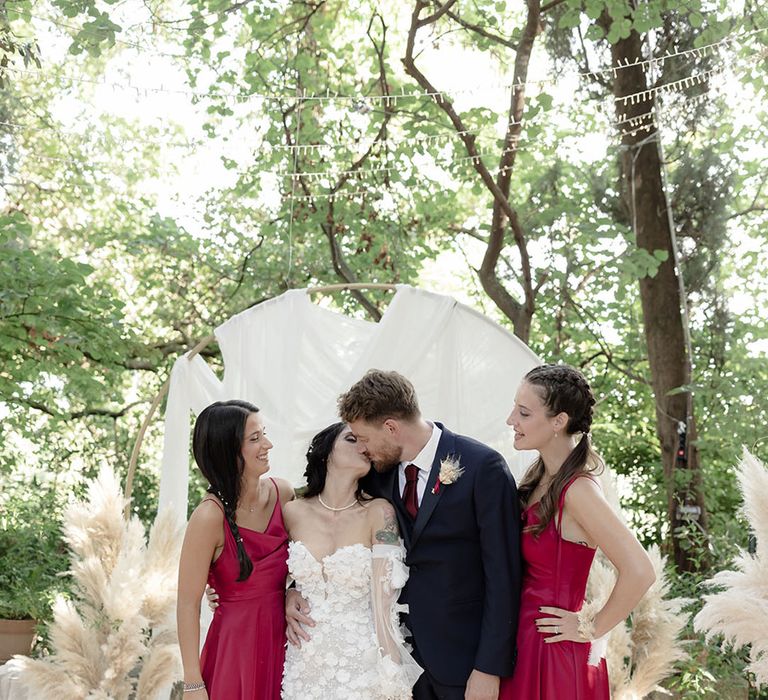 Bridesmaids wear satin burgundy bridesmaid dresses whilst stood beside bride & groom during destination wedding 