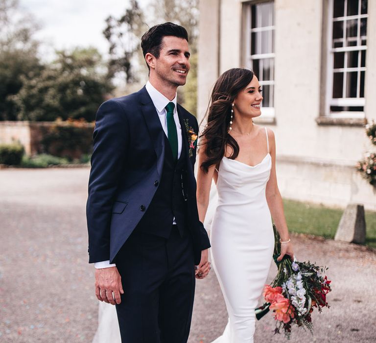 Bride with pearl earrings in Kyha wedding dress with a cowl neck and open back walking with the groom in a three piece navy suit 