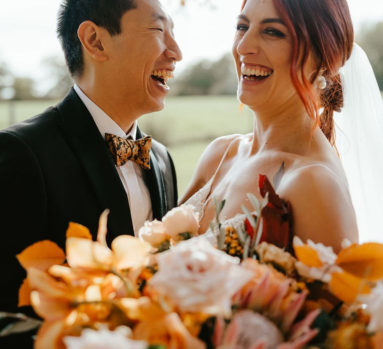 Groom laughs beside his bride who holds large orange bridal bouquet 