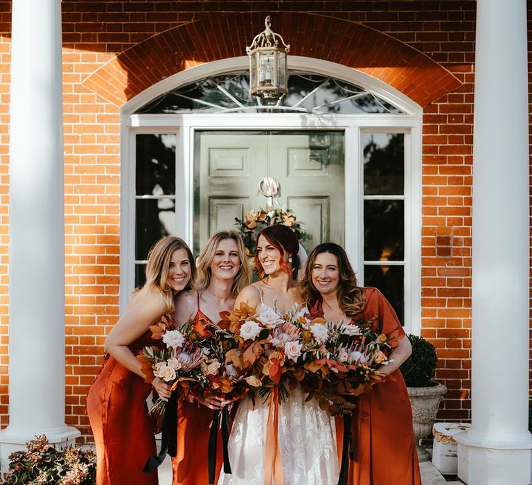 Bride stands with her bridesmaids holding orange bouquets and wearing satin orange bridesmaid dresses 