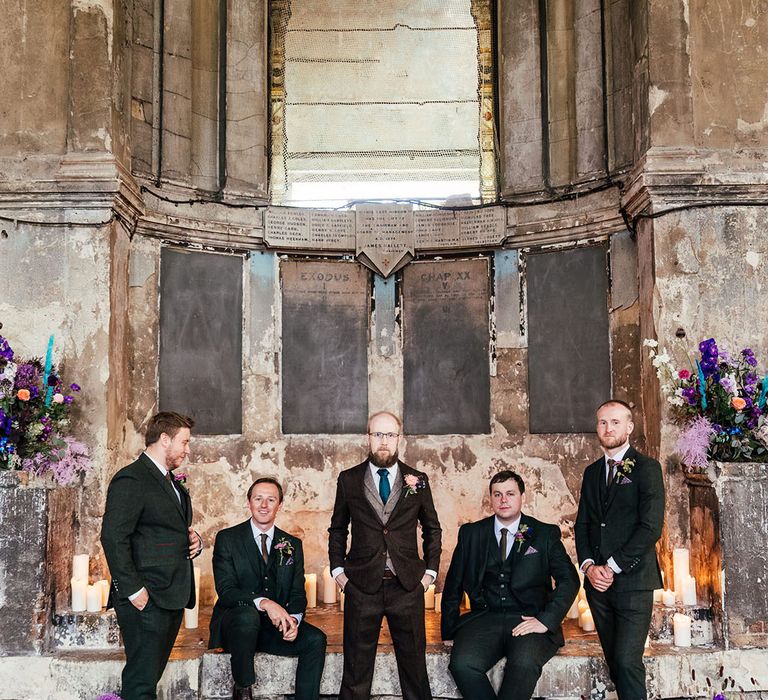 Groom in mocha brown textured suit with deep green tie and pink and purple boutonniere standing at the alter of Asylum Chapel London with groomsmen wearing dark three piece suits with dried flower boutonniere 