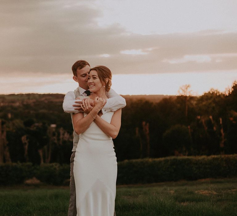 Groom wraps his arms around bride in Savannah Miller wedding dress during golden hour portraits