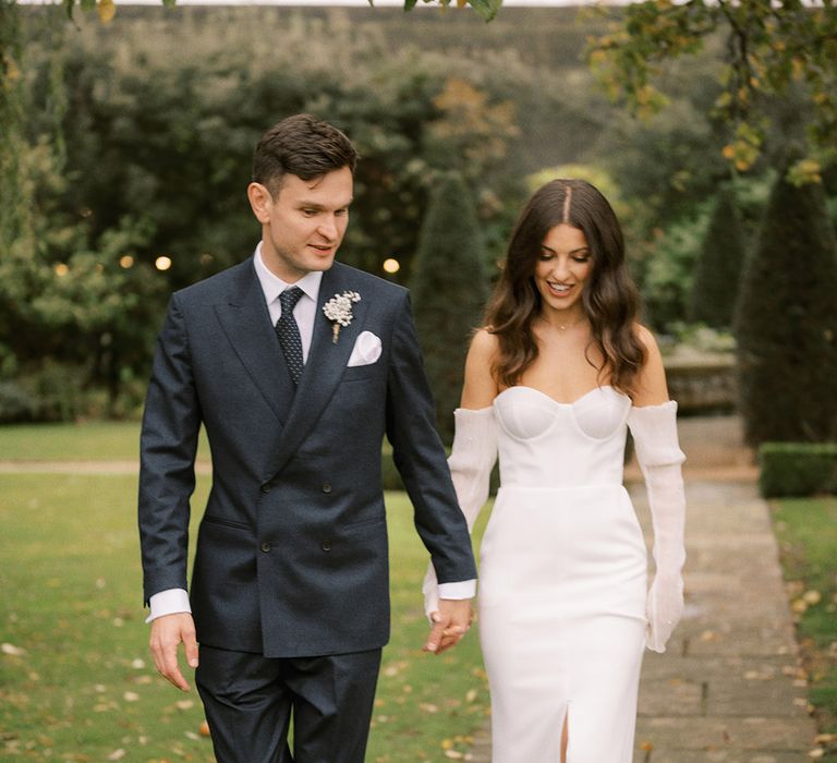 Groom in double breasted suit jacket walking hand in hand with the bride in a fitted wedding dress with detachable sleeves with pearls 