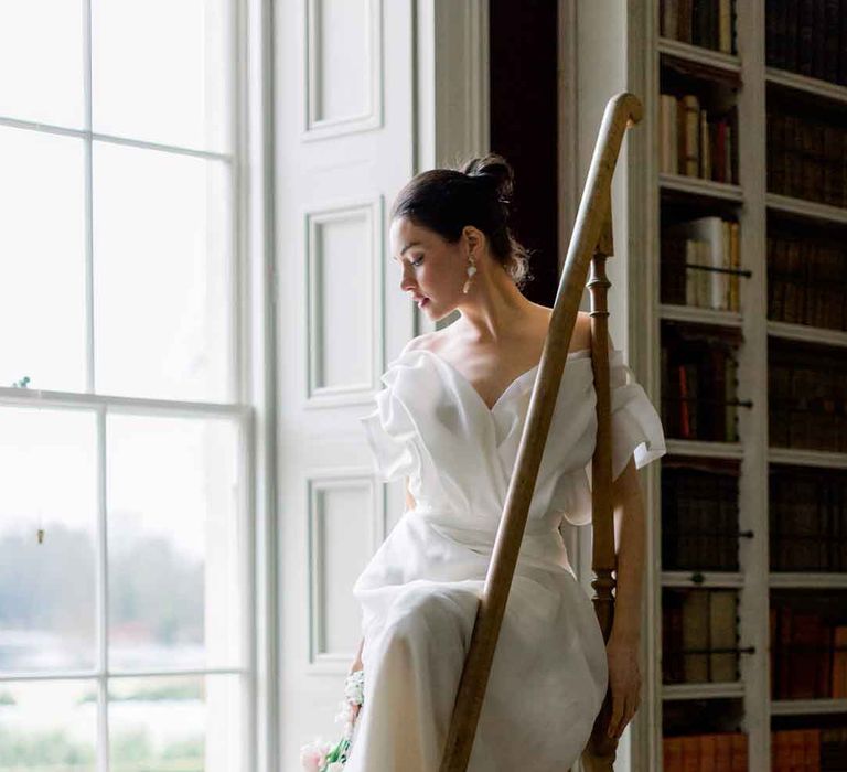 Bride wearing sculpted, layered off the shoulder wedding dress with open toe wedding heels sitting on ladder at St Giles House 